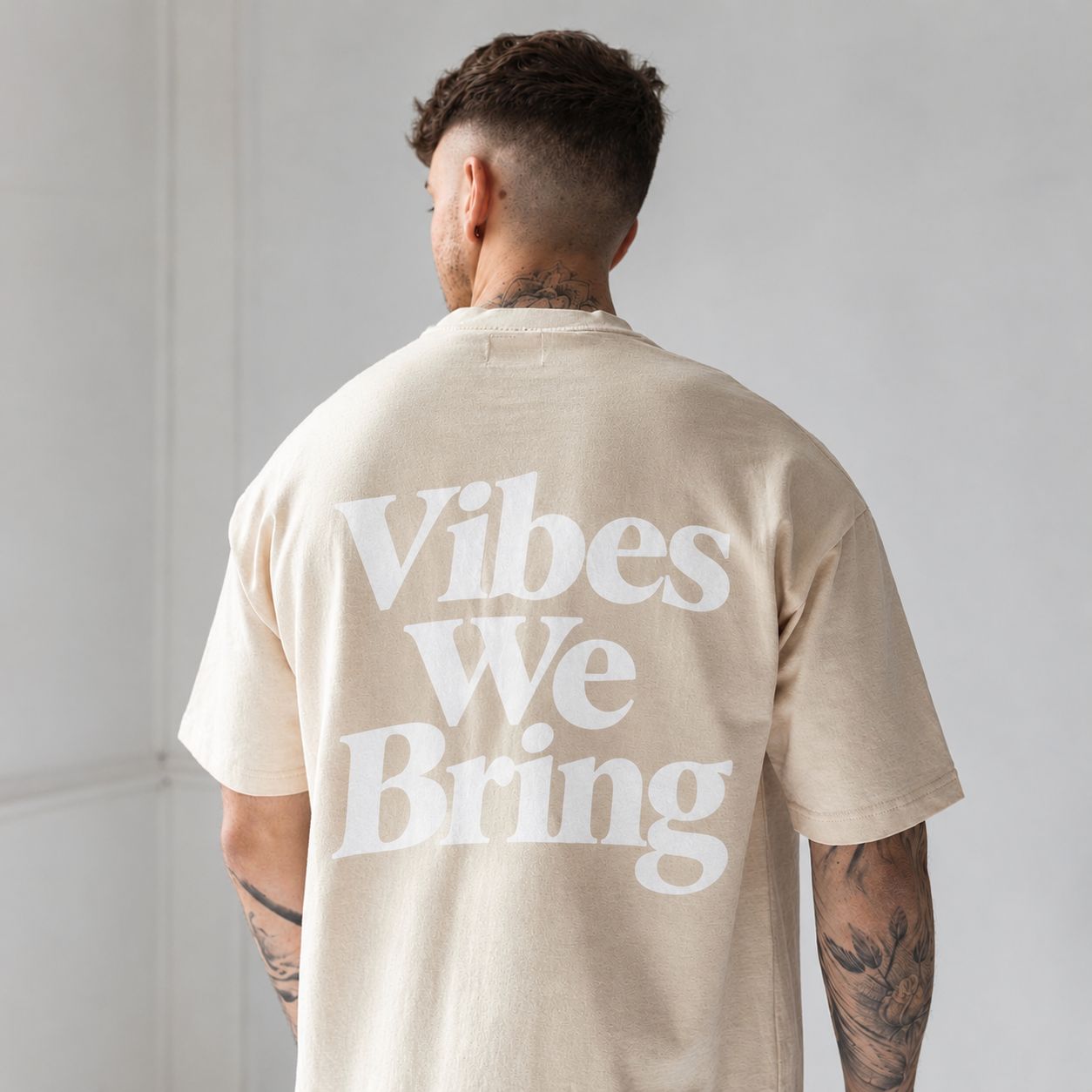 A man photographed from behind in an oversized cream T-shirt with Vibes We Bring printed across the back in large white serif lettering.