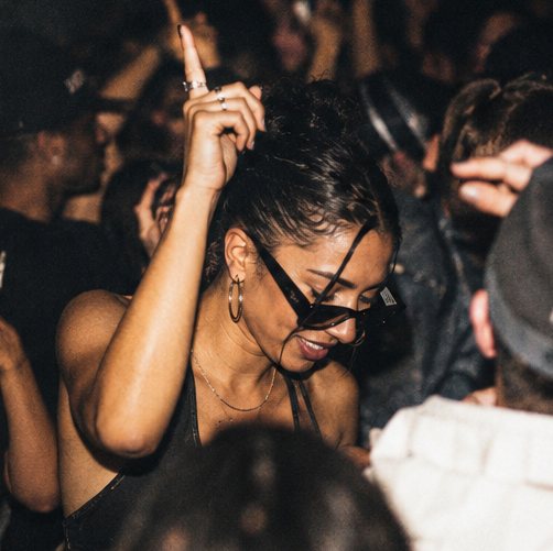 A woman in sunglasses dancing with one hand raised in a packed crowd.