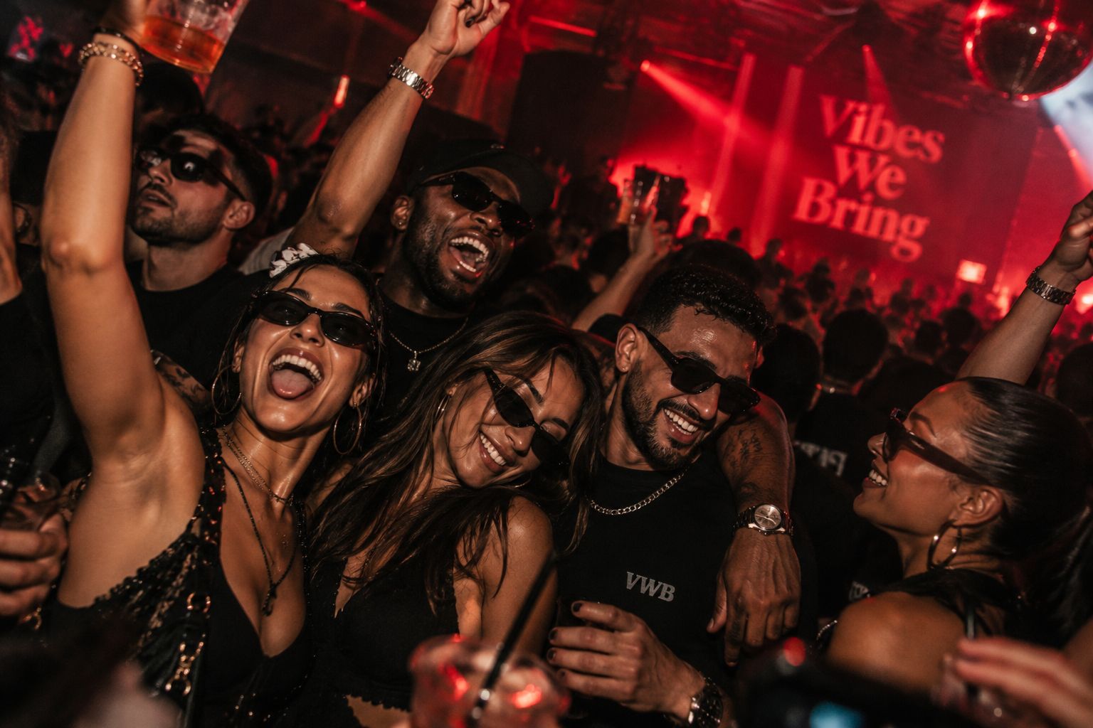 A group of friends in sunglasses laughing and dancing close together in a red-lit crowd, one woman with an arm raised, with a large serif Vibes We Bring screen glowing behind them.