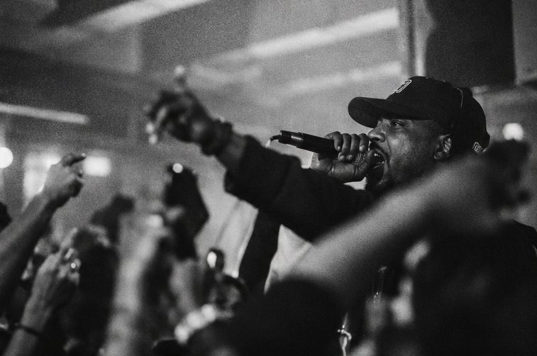Black-and-white photograph of an MC in a cap with a microphone, surrounded by raised hands.