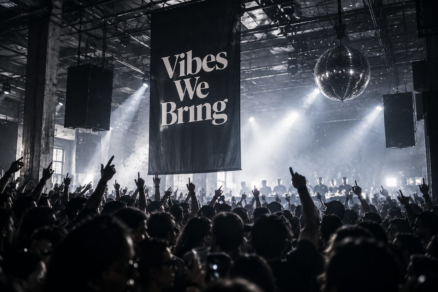 Black-and-white view from inside a packed crowd, hands raised and pointing upwards, towards a tall black Vibes We Bring banner, a mirror ball and a row of DJs at the back of a large industrial hall.